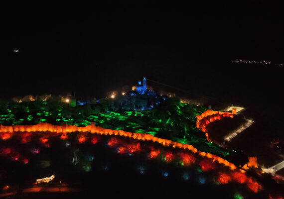 Дрон снимка от високо на нощния спектакъл 'Звук и светлина' във Велико Търново, показваща крепостта Царевец, осветена в червено, зелено и синьо, със стотици хора, събрани долу.