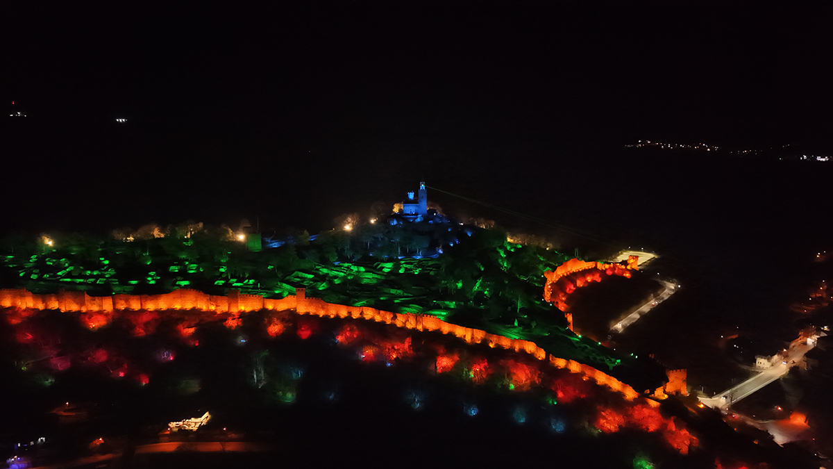 Дрон снимка от високо на нощния спектакъл 'Звук и светлина' във Велико Търново, показваща крепостта Царевец, осветена в червено, зелено и синьо, със стотици хора, събрани долу.