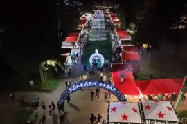 Дрон кадър на Коледен базар във Велико Търново през нощта, показващ осветените къщички, арката "КОЛЕДЕН БАЗАР" и централния светлинен декор.